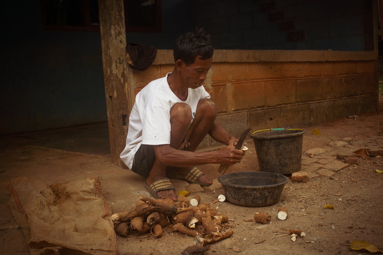 Man sitting on the ground peeling cassava roots with a knife, depicting traditional lifestyle.